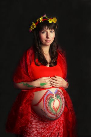 Bohyně země (Očekávání) – barevná fotografie, žena v červených šatech, má na hlavě věneček z květin a ukazuje těhotenské bříško, pomalované barvami