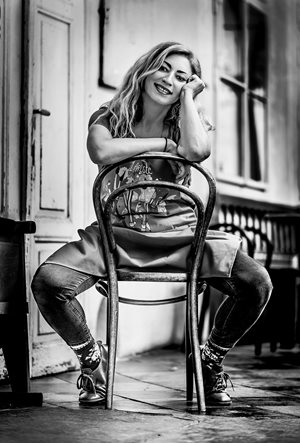 Iva – černobílá fotografie, žena sedí obkročmo na staré židli z ohýbaného dřeva, o jejíž opěradlo se opírá jednou rukou, druhou si podepírá hlavu