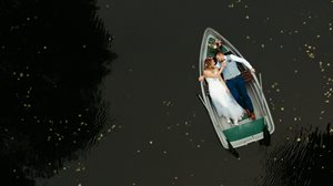 Loďka – barevná fotografie pořízená z výšky, na vodní hladině pluje loďka, v ní leží muž a žena ve svatebním oblečení, novomanželé